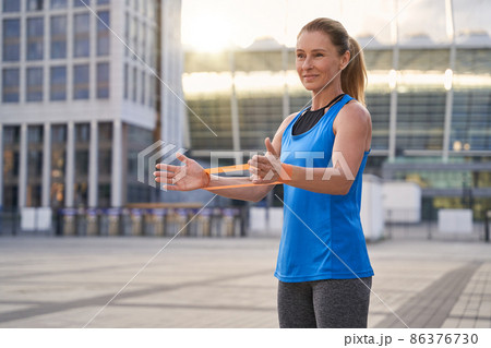 Beautiful sportive woman in sportswear, female trainer holding resistance band while standing outdoors, working out in the city Beautiful sportive woman in sportswear, female trainer holding resistance band while standing outdoors, working out in the city 86376730