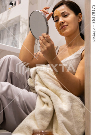 Smiling woman applying scrub peeling face mask massaging skin looking at mirror reflection bathroom Smiling woman applying scrub peeling face mask massaging skin looking at mirror reflection bathroom 86379175