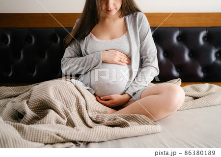 Pregnant woman hugs belly with hands while sitting on bed Pregnant woman hugs belly with hands while sitting on bed 86380189