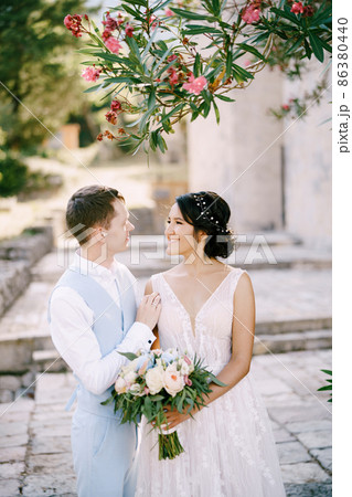 The bride and groom are embracing under a blooming oleander, the bride is holding a bouquet in her hands  86380440