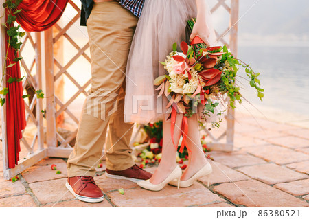 Groom and bride with a bouquet of flowers stand on a stone tile against the background of a carved screen decorated with a red curtain and greenery. Close-up Groom and bride with a bouquet of flowers stand on a stone tile against the background of a carved screen decorated with a red curtain and greenery. Close-up 86380521