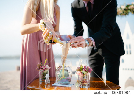 Groom and bride pour multi-colored sand in layers into a glass jar 86381004