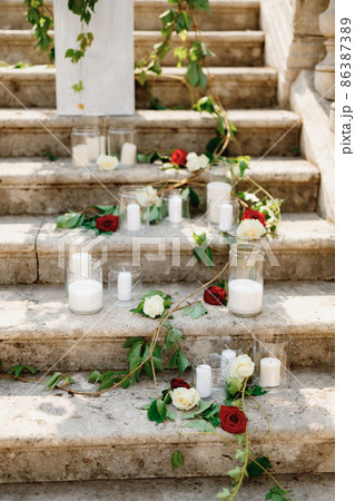 White candles in glass jars decorated with roses and green branches on stone steps 86387389