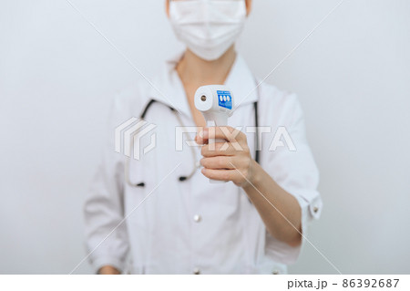 Temperature measurement gun in doctor hands. Close-up shot of doctor wearing protective surgical mask ready to use infrared isometric thermometer gun to check body temperature for virus symptoms. 86392687