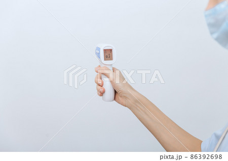 Temperature measurement gun in doctor hands. Close-up shot of doctor wearing protective surgical mask ready to use infrared isometric thermometer gun to check body temperature for virus symptoms. Temperature measurement gun in doctor hands. Close-up shot of doctor wearing protective surgical mask ready to use infrared isometric thermometer gun to check body temperature for virus symptoms. 86392698