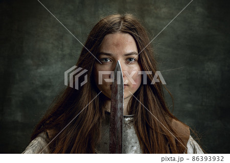 Vintage portrait of adorable woman, medieval female warrior or knight with dirty wounded face looking at camera isolated over dark retro background. 86393932