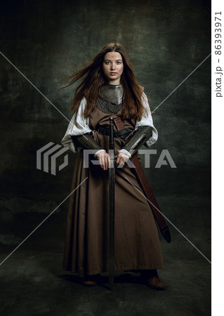 Full-length portrait of seriuos beautiful woman in image of medieval warrior or knight with dirty wounded face with big sword isolated over dark vintage background. 86393971