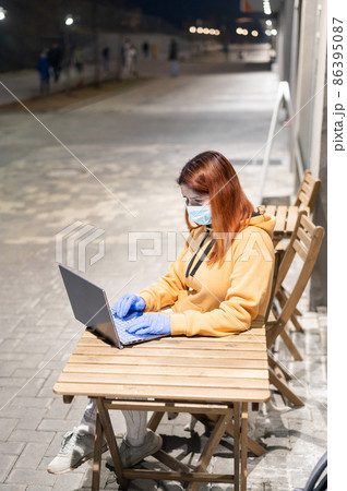 A woman in a medical mask and gloves remotely works late at night outdoors. Ocher-colored hoodie girl with laptop on the street during quarantine. Coronavirus. 86395087