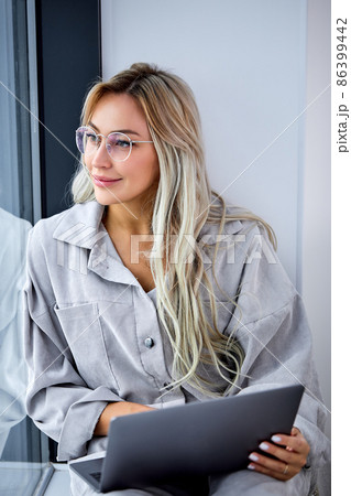 blonde female in casual clothing using computer laptop and sitting on window sill blonde female in casual clothing using computer laptop and sitting on window sill 86399442