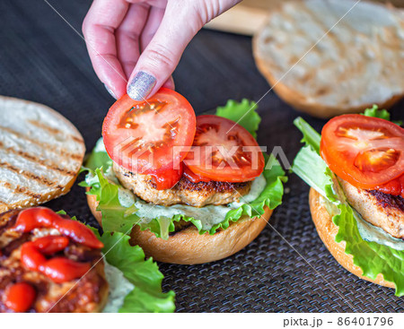 A woman's hand lays down a slice of tomato on a delicious homemade burger. Handmade homemade burgers, close-up A woman's hand lays down a slice of tomato on a delicious homemade burger. Handmade homemade burgers, close-up 86401796