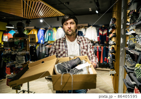 Salesman advises client on choice of hiking shoes in an outdoor store. Male tourist shop owner helping customer try on hiking boots. Young seller confidently presents mountain boot. sell, trade Salesman advises client on choice of hiking shoes in an outdoor store. Male tourist shop owner helping customer try on hiking boots. Young seller confidently presents mountain boot. sell, trade 86405595