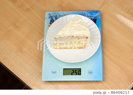 A piece of delicious cake with white butter cream lies on the scales. Calorie counting concept, figure tracking A piece of delicious cake with white butter cream lies on the scales. Calorie counting concept, figure tracking 86406217