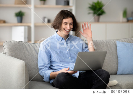 Distant communication concept. Happy man video calling on laptop computer, waving hand to screen, sitting on sofa 86407098