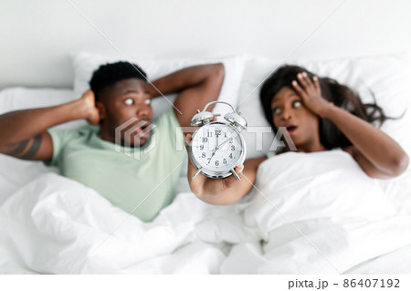 Shocked millennial black male and female with open mouths show alarm clock, lies on white bed in bedroom 86407192