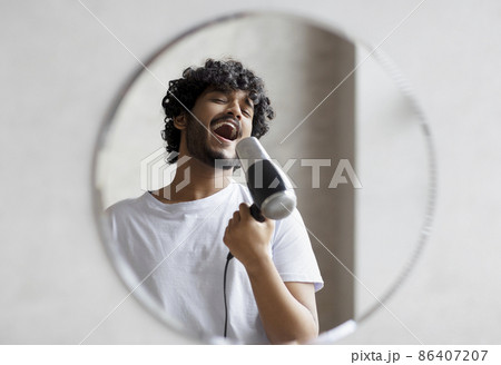 Joyful indian man having fun in the morning in bathroom, holding hairdryer and singing as in microphone Joyful indian man having fun in the morning in bathroom, holding hairdryer and singing as in microphone 86407207