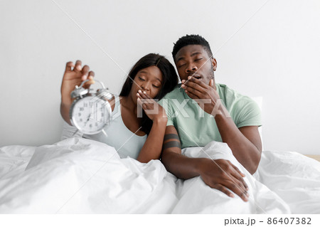 Tired sad sleepy millennial african american couple sit on white bed in bedroom, yawn and show alarm clock 86407382