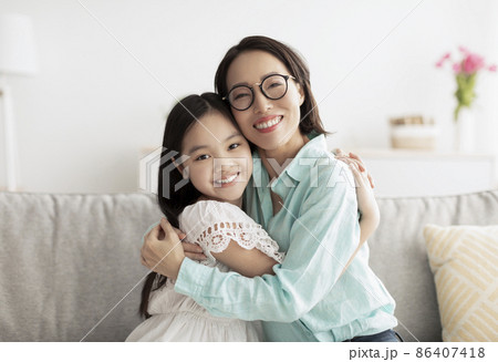 Happy little Asian girl and her loving grandmother hugging and looking at camera on couch at home 86407418