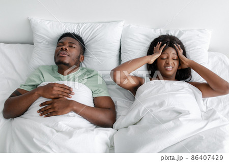 Sad despaired young african american wife suffering from headache, noise and snoring sleeping husband on bed Sad despaired young african american wife suffering from headache, noise and snoring sleeping husband on bed 86407429