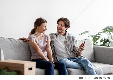 Pleased caucasian millennial husband show tablet to wife on sofa in living room with cardboard boxes Pleased caucasian millennial husband show tablet to wife on sofa in living room with cardboard boxes 86407576
