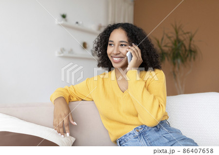 Cheery young African American woman talking on smartphone, sitting on sofa at home Cheery young African American woman talking on smartphone, sitting on sofa at home 86407658