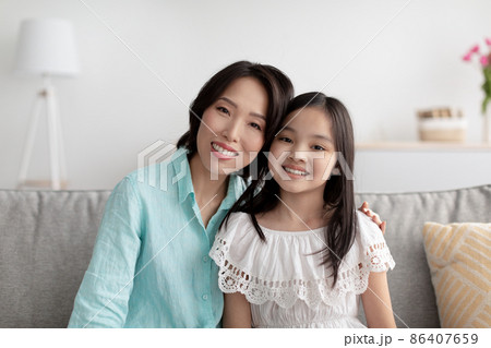 Portrait of cheerful mature Asian woman and her adorable granddaughter sitting on couch at home, smiling at camera 86407659