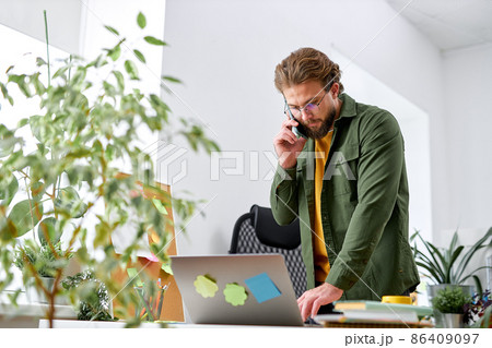 young bearded man standing behind table and talking on phone. Businessman is serious young bearded man standing behind table and talking on phone. Businessman is serious 86409097