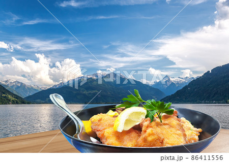 Traditional austrian food (Wiener schnitzel) against Zell am see village with lake in Austria 86411556