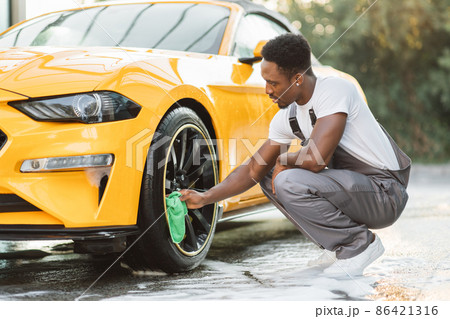 Handsome African guy wiping wheel of modern luxury car with green microfiber cloth 86421316