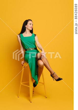 Cute woman in green dress sitting over yellow studio background Cute woman in green dress sitting over yellow studio background 86426089