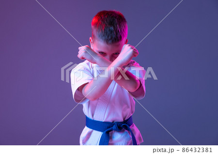 Dynamic portrait of kid, young male taekwondo, karate athletes in doboks doing basic movements isolated on purple background in neon. Concept of sport, martial arts 86432381