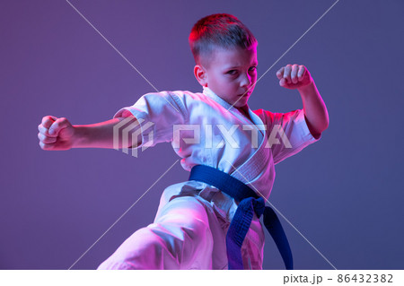 Dynamic portrait of kid, young male taekwondo, karate athletes in doboks doing basic movements isolated on purple background in neon. Concept of sport, martial arts 86432382