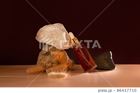Transparent glass cosmetic dropper standing near stones and dry flowers on dark red and trendy beige background. Beauty composition. Concept of natural and original skincare products presentation.  86437710