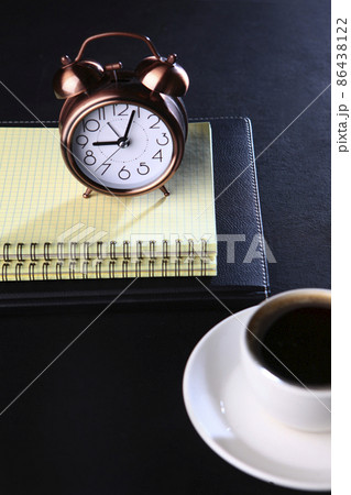 Alarm clock and notepad for notes.A cup of coffee is out of focus.Food and drinks.Coffee break. Space for copying.Black background.Vertical photo. 86438122