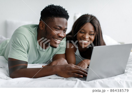 Laughing millennial black guy and woman looking at computer lie on white bed in bedroom Laughing millennial black guy and woman looking at computer lie on white bed in bedroom 86438579