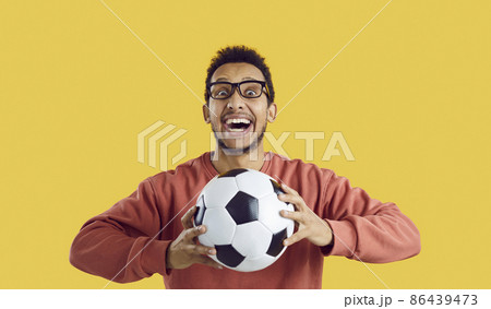 Crazy soccer fan supporting favorite team. Portrait of happy funny excited cheerful black man standing isolated on solid yellow colour background, holding football, looking at camera and smiling 86439473