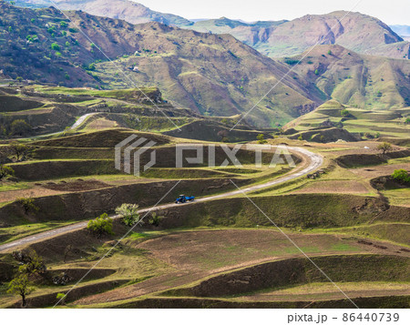 Terraces of spring mountain agriculture. Old tractor is driving along a winding mountain dirt road. Dangerous narrow mountain road through the hills to a high-altitude village. Terraces of spring mountain agriculture. Old tractor is driving along a winding mountain dirt road. Dangerous narrow mountain road through the hills to a high-altitude village. 86440739