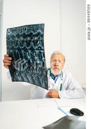Vertical shot of serious mature adult male doctor holding MRI spine image of patient sitting at desk in medical office room, looking at camera, thinking about diagnosis, expressing concern of illness. Vertical shot of serious mature adult male doctor holding MRI spine image of patient sitting at desk in medical office room, looking at camera, thinking about diagnosis, expressing concern of illness. 86449569