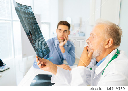 Thoughtful mature adult male physician consult frustrated young man patient giving bad news explaining results of MRI image. Unhappy scared young man listening to bad news sitting in doctor office. Thoughtful mature adult male physician consult frustrated young man patient giving bad news explaining results of MRI image. Unhappy scared young man listening to bad news sitting in doctor office. 86449570