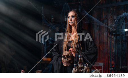 Halloween concept. Witch portrait close up with dreadlocks looking away dressed black hood standing dark room with cage on background 86464189