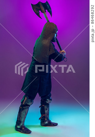Studio shot of young bearded man in image of medieval warrior, archery in chain armor with ax isolated over purple background in neon. Comparison of eras 86468292