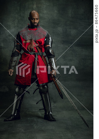 Vintage style portrait of brutal dark skinned man, medieval warrior or knight with wounded face wearing armour isolated over dark background. Comparison of eras, history Vintage style portrait of brutal dark skinned man, medieval warrior or knight with wounded face wearing armour isolated over dark background. Comparison of eras, history 86475660
