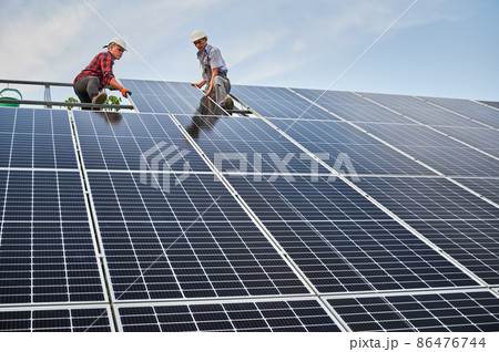Low angle of two men solar technicians building photovoltaic solar panels under blue sky. Male workers in safety helmets placing solar module on metal rails. Concept of alternative energy sources. 86476744