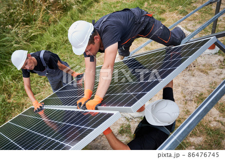 Male workers building photovoltaic solar panel system outdoors. Men engineers placing solar module on metal rails, wearing construction helmets and work gloves. Renewable and ecological energy. 86476745