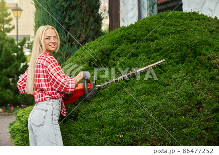 Young woman pruning bushes with electric hedge trimmer 86477252