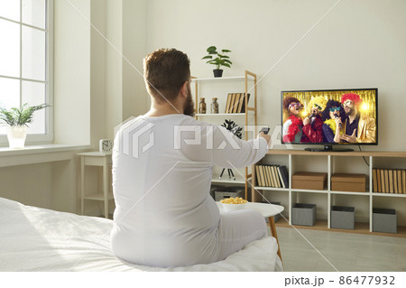 Plump ginger guy wakes up in morning and switches television with remote control. Funny fat man in PJs sitting in bedroom at home and watching entertainment channel on widescreen HD LED TV, back view 86477932