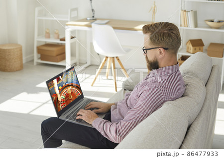 Young man sit relax on sofa play casino online on computer. Caucasian male rest at home couch gamble internet on laptop. Entertainment on web. Gambler or gamer Russian roulette. 86477933
