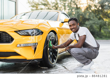 Young guy in gray overalls African bearded man cleaning a wheel, car rims Young guy in gray overalls African bearded man cleaning a wheel, car rims 86481281
