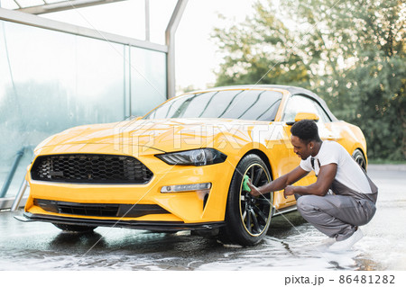 Handsome African guy wiping with green cloth car wheel during the washing process outdoors. Handsome African guy wiping with green cloth car wheel during the washing process outdoors. 86481282