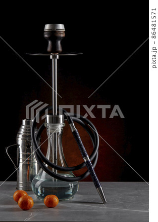 Still life with a beautiful modern hookah standing on the table, with a metal protective cap and three ripe juicy tangerines, on a dark red background. Space for copying. Vertical location. Still life with a beautiful modern hookah standing on the table, with a metal protective cap and three ripe juicy tangerines, on a dark red background. Space for copying. Vertical location. 86481571