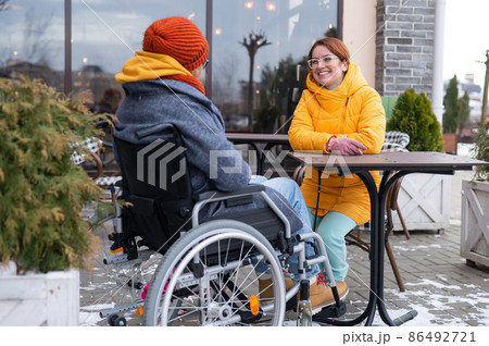 Two girlfriends in a cafe on a street terrace in winter. Woman in a wheelchair. Two girlfriends in a cafe on a street terrace in winter. Woman in a wheelchair. 86492721